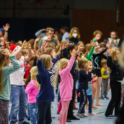 Gruppierung von Kindern mit erhobenen Händen in einer Sporthalle, begleitet von Erwachsenen im Hintergrund.