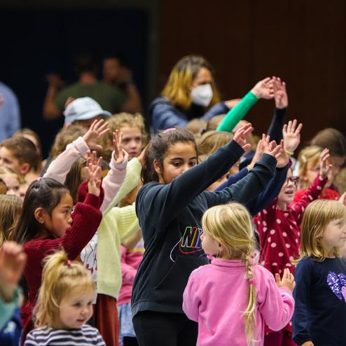 Eine Gruppe von Kindern mit erhobenen Händen tanzt fröhlich in einer Halle.