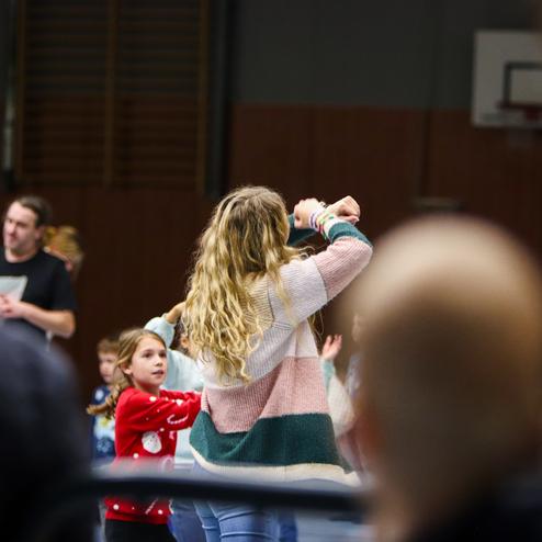 Eine Gruppe von Kindern und Erwachsenen tanzt auf einem Sportplatz, verschiedene Aktivitäten und Freude sind sichtbar.