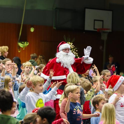 Weihnachtsfeier mit Kindern, die Freude zeigen, während ein verkleideter Weihnachtsmann im Vordergrund tanzt und winkt.