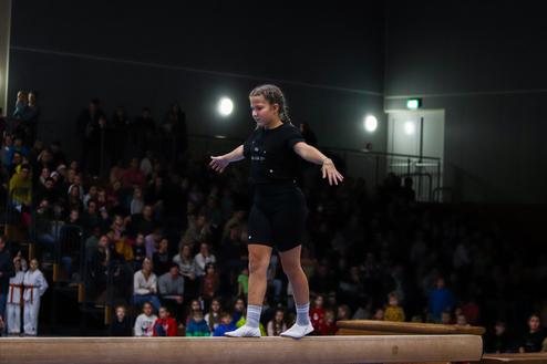Junge Sportlerin balanciert auf einem Schwebebalken, während eine große Menge im Hintergrund zuschaut.