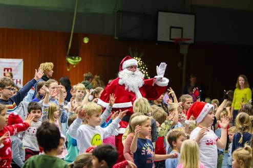 Weihnachtsfeier mit Kindern, die Freude zeigen, während ein verkleideter Weihnachtsmann im Vordergrund tanzt und winkt.