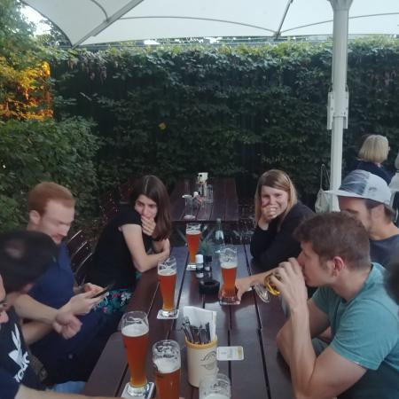 Gruppe von Menschen sitzt an einem Tisch im Freien, umgeben von Pflanzen, mit Getränken vor sich.