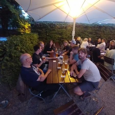 Gruppe von Personen sitzt an einem Tisch unter einem Sonnenschirm, umgeben von grünen Pflanzen, bei Abendlicht.