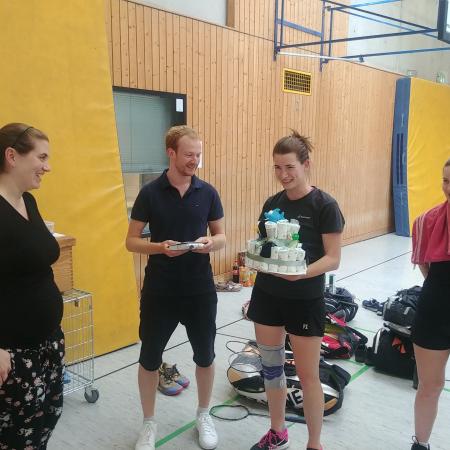 Vier Personen in Sportkleidung stehen in einer Turnhalle und feiern mit einer Torte und Geschenken.