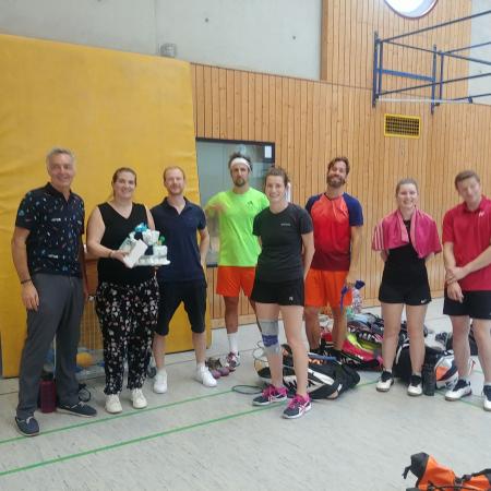 Gruppe von sechs Personen in Sportkleidung, posierend mit Trophäe in einer Sporthalle. Sportgeräte liegen auf dem Boden.