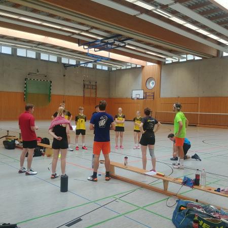Gruppentraining in einer Sporthalle mit sieben Personen in Sportkleidung, Badminton-Equipment und einer Wand mit Uhr.