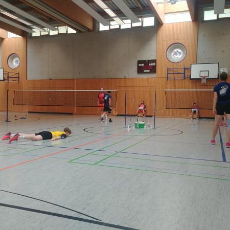 Badminton-Spieler in einer Halle, einige Spieler aktiv, einer am Boden liegend, Badmintonnnetze im Hintergrund sichtbar.