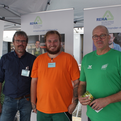 Drei M&auml;nner stehen l&auml;chelnd vor Informationsst&auml;nden der REHA, jeder tr&auml;gt ein Namensschild und verschiedene T-Shirts.