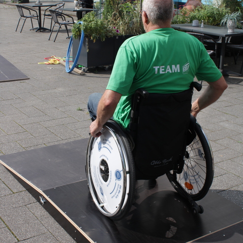 Mann im Rollstuhl f&auml;hrt auf einer Rampe, umgeben von Pflanzen und Tischen in einem offenen Platz.