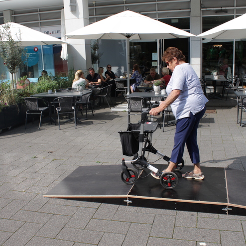 Eine &auml;ltere Frau geht mit einem Rollator &uuml;ber eine ramponierte Holzplanke vor einem Caf&eacute; im Freien.