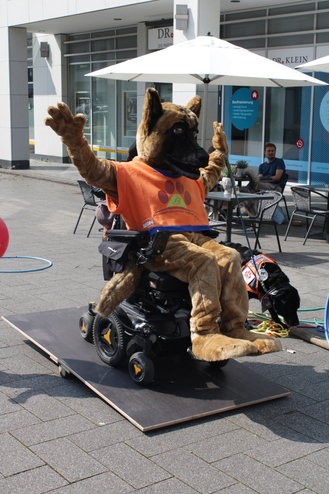 Mensch in einem Hundekostüm sitzt in einem Elektrorollstuhl auf einer Bühne, während ein Assistenzhund neben ihm sitzt.