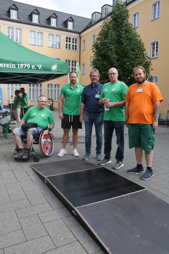 Fünf Personen stehen neben einer mobilen Rampe auf einem Platz, umgeben von Stühlen und einem grünen Pavillon.