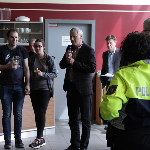 Ein Mann spricht mit Mikrofon, w&auml;hrend Paare mit Getr&auml;nken und Polizisten in einem Raum stehen.