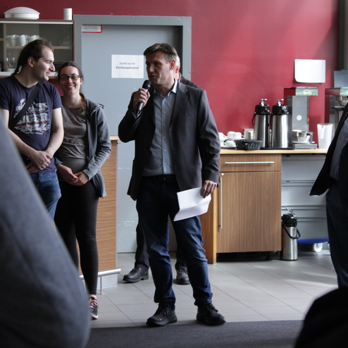 Mann spricht mit Mikrofon vor Publikum in einer Cafeteria, w&auml;hrend zwei Personen ihm aufmerksam zuh&ouml;ren.