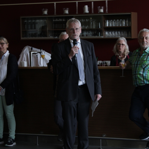 Ein Mann in Anzug h&auml;lt ein Mikrofon und spricht, w&auml;hrend mehrere Personen im Hintergrund zuh&ouml;ren.