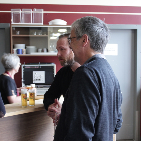 Zwei M&auml;nner unterhalten sich an einem Tisch, im Hintergrund ist eine Frau beim Servieren von Getr&auml;nken zu sehen.