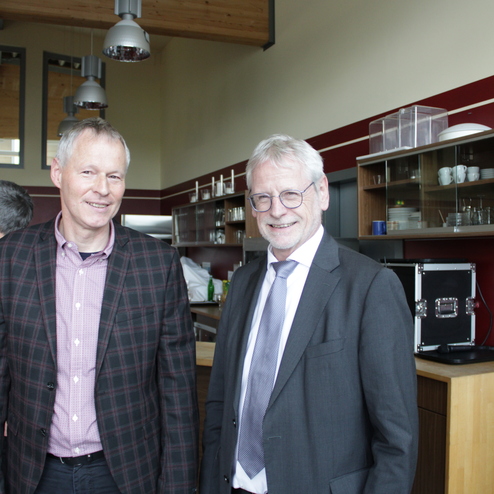 Zwei M&auml;nner stehen l&auml;chelnd in einer modernen Umgebung mit Holz- und Glaselementen, w&auml;hrend sich im Hintergrund weitere Personen unterhalten.