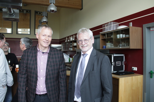 Zwei Männer stehen lächelnd in einer modernen Umgebung mit Holz- und Glaselementen, während sich im Hintergrund weitere Personen unterhalten.