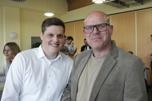 Zwei Männer lächeln in einer hellen Umgebung, während im Hintergrund weitere Personen in Gesprächen zu sehen sind.