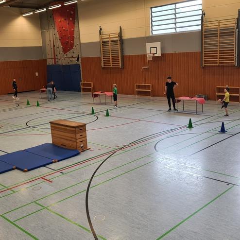 Sporthalle mit Hindernissen: Kinder springen über Matten und Kisten, während Erwachsene hilfreiche Anweisungen geben.