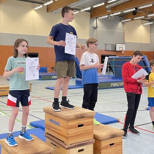 Fünf Kinder mit Urkunden stehen auf Holzpodesten in einer Sporthalle. Geräte im Hintergrund sichtbar.