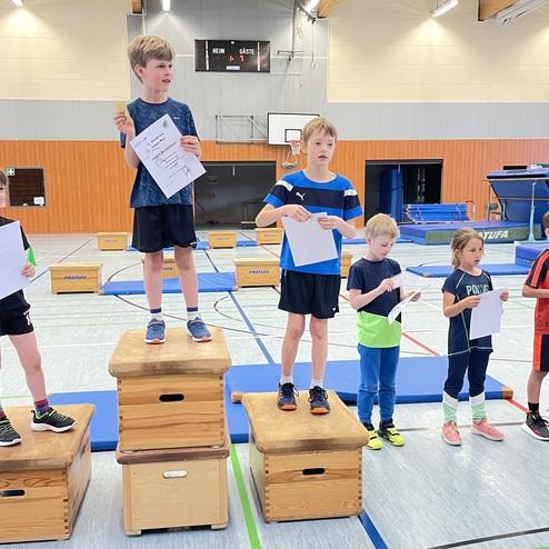 Fünf Kinder stehen auf Holzkisten und halten Urkunden in der Sporthalle, während der Sieger eine Medaille präsentiert.