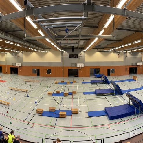 Blick in eine Sporthalle mit Turnger&auml;ten, Matten und Sch&uuml;lern, die sich auf verschiedenen Anlagen bewegen.