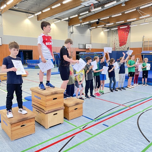 Gruppe von Kindern steht auf Holzbl&ouml;cken in einer Turnhalle und h&auml;lt Urkunden in die H&ouml;he.