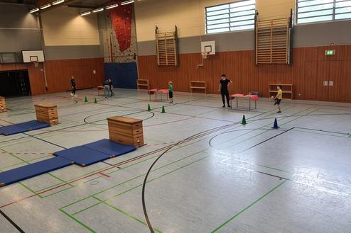 Sporthalle mit Hindernissen: Kinder springen über Matten und Kisten, während Erwachsene hilfreiche Anweisungen geben.