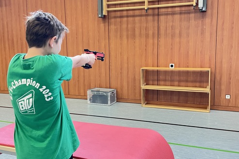 Junge in grüner T-Shirt zielt mit einem Spielzeugpistole auf eine Wand in einer Turnhalle. Pinke Matte auf einem Tisch.
