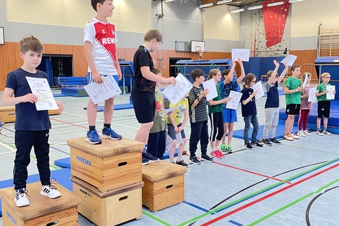 Gruppe von Kindern steht auf Holzblöcken in einer Turnhalle und hält Urkunden in die Höhe.
