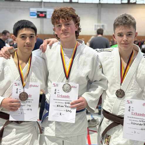 Drei Jugendliche in Judoanz&uuml;gen halten Medaillen und Urkunden bei einem Wettkampf in einer Sporthalle.