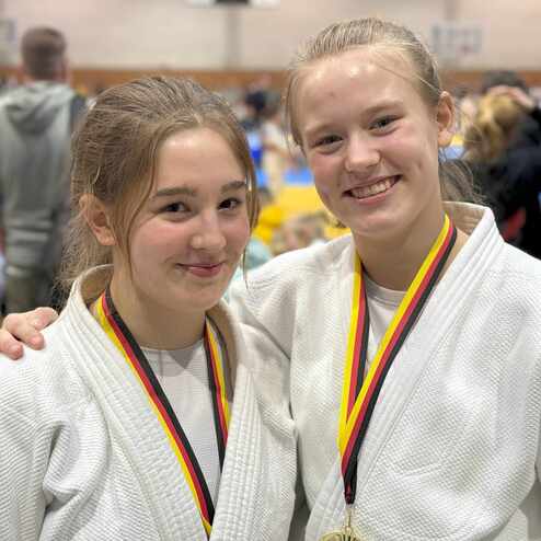 Zwei junge Judoka in wei&szlig;en Anz&uuml;gen mit Medaillen l&auml;cheln vor einer Wettkampfumgebung.