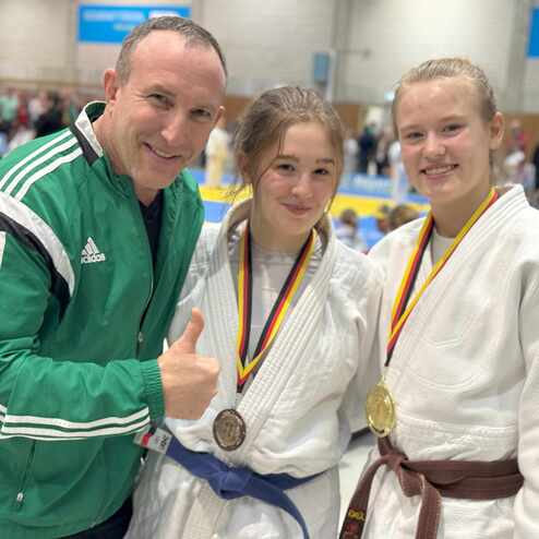 Drei Personen l&auml;cheln zusammen, zwei in Judogis mit Medaillen, eine Person gibt Daumen hoch im Hintergrund einer Wettkampfst&auml;tte.