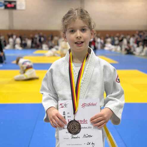 M&auml;dchen in Judogi mit Medaille und Urkunde, auf einer Judo-Matte, im Hintergrund Wettk&auml;mpfer und Zuschauer.