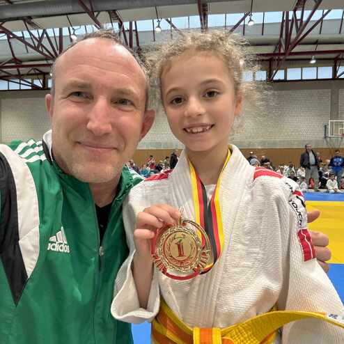 Junge mit lockigen Haaren und Judoanzug h&auml;lt stolz eine Medaille, w&auml;hrend ein Trainer in Sportjacke neben ihr posiert.