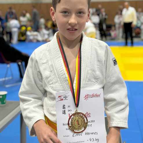 Junge in Judo-Gi mit Medaille und Urkunde f&uuml;r den ersten Platz bei einem Wettkampf, im Hintergrund weitere Judo-Teilnehmer.