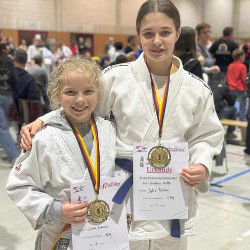 Zwei M&auml;dchen in Judowesten halten Urkunden und Medaillen bei einem Wettbewerb in einer Sporthalle.