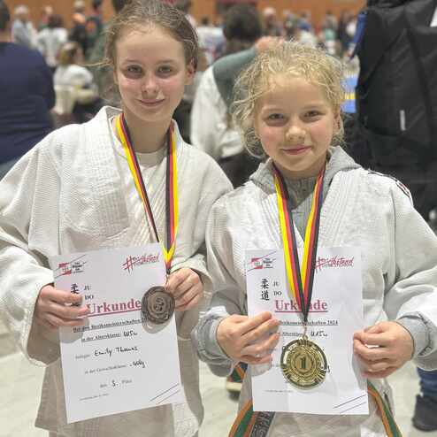 Zwei M&auml;dchen in Judoanz&uuml;gen halten Urkunden und Medaillen auf einem Wettkampf im Hintergrund.