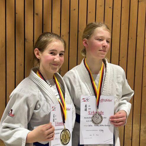 Zwei M&auml;dchen in Judokleidung halten Medaillen und Urkunden vor einer Holzverkleidung nach einem Wettkampf.