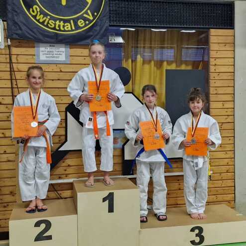 Vier junge Judoka stehen auf dem Podest mit Medaillen und Urkunden, begleitet von einem Trainer im Hintergrund.