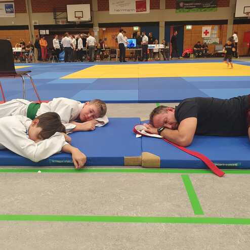 Drei Personen in Judokleidung und Sportbekleidung ruhen auf blauen Matten in einer Sporthalle.