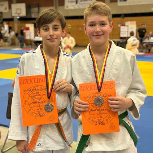 Zwei Jungen in Judogis halten Urkunden für den 3. Platz bei einer Bezirksmeisterschaft in der Sporthalle.