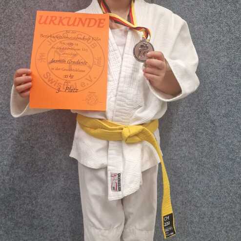 Mädchen in Judokleidung hält eine Urkunde und eine Medaille vor einer grauen Wand.