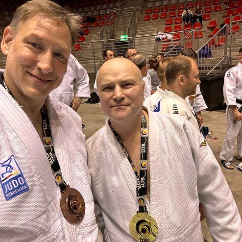 Zwei Judokas stehen smiling in einem Turnierumfeld, tragen Medaillen und Judoanz&uuml;ge vor einer Zuschauertrib&uuml;ne.