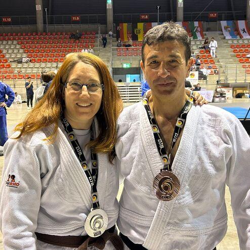 Paar in Judogis mit Medaillen l&auml;chelt vor einem Sport-Event-Hintergrund mit leeren Sitzen und anderen K&auml;mpfern.