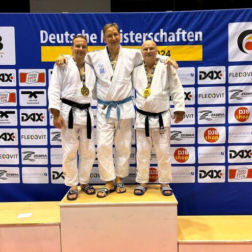 Drei Judoka mit Medaillen stehen auf einem Podest vor einem Banner der Deutschen Meisterschaften 2024.