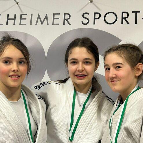 Drei Jugendliche in Judokas mit Medaillen lächeln vor einem Sportverein-Hintergrund.