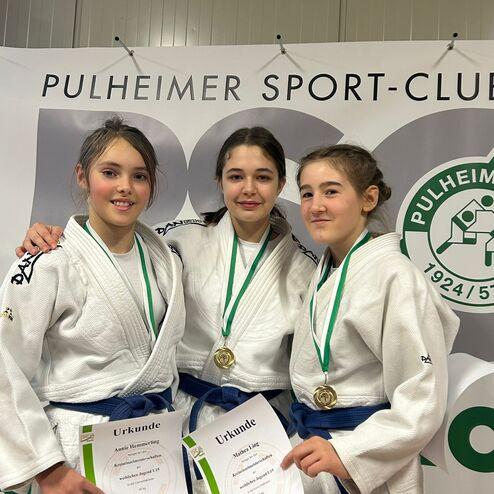 Drei Mädchen in Judogewändern halten Urkunden und Medaillen, vor einem Banner des Pulheimer Sport-Clubs.